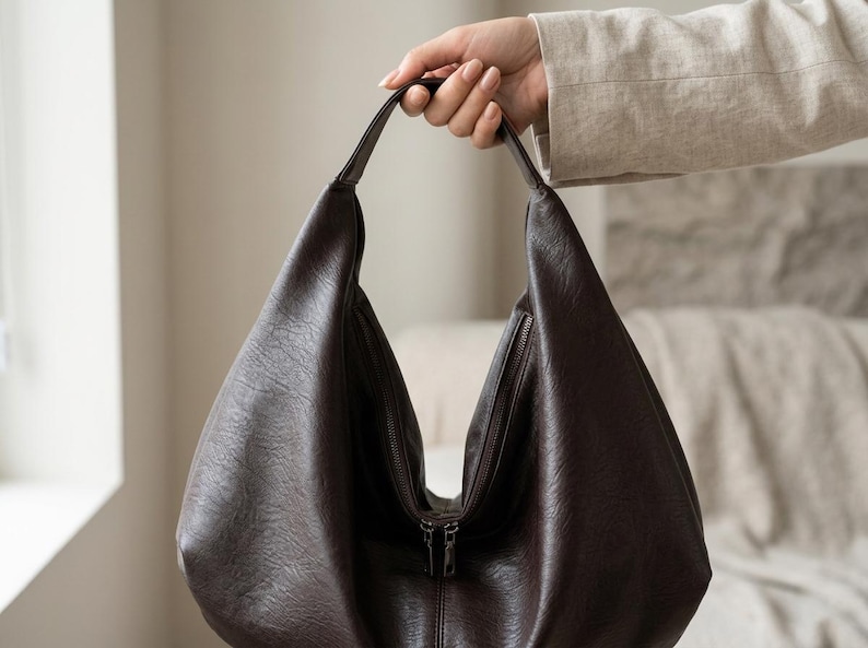 Op de afbeelding: Een donkerbruine hobo-tas met een ritssluiting. De tas is gemaakt van een textuurmateriaal en heeft een enkele handgreep. De tas wordt door een hand omhoog gehouden.
