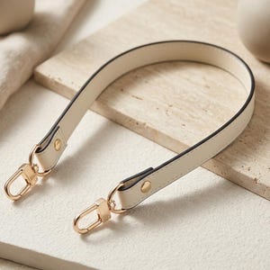 Peut inclure: Bandoulière de sac à main crème avec des accessoires dorés. La sangle est en similicuir lisse avec des bordures noires. Les fermoirs dorés sont fixés à la sangle avec de petits rivets dorés. La sangle est incurvée et repose sur une surface claire.