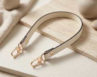 Op de afbeelding: Crèmekleurige handtasriem met goudkleurige hardware. De riem is gemaakt van glad imitatieleer met zwarte randen. De gouden sluitingen zijn met kleine gouden klinknagels aan de riem bevestigd. De riem is gebogen en ligt op een lichtgekleurd oppervlak.