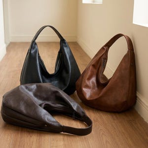 Op de afbeelding: Drie leren hobo-tassen in zwart, bruin en donkerbruin worden getoond op een houten vloer. De tassen hebben een halvemaanvorm, een enkele schouderriem en een ritssluiting. De tassen zijn leeg en staan rechtop.