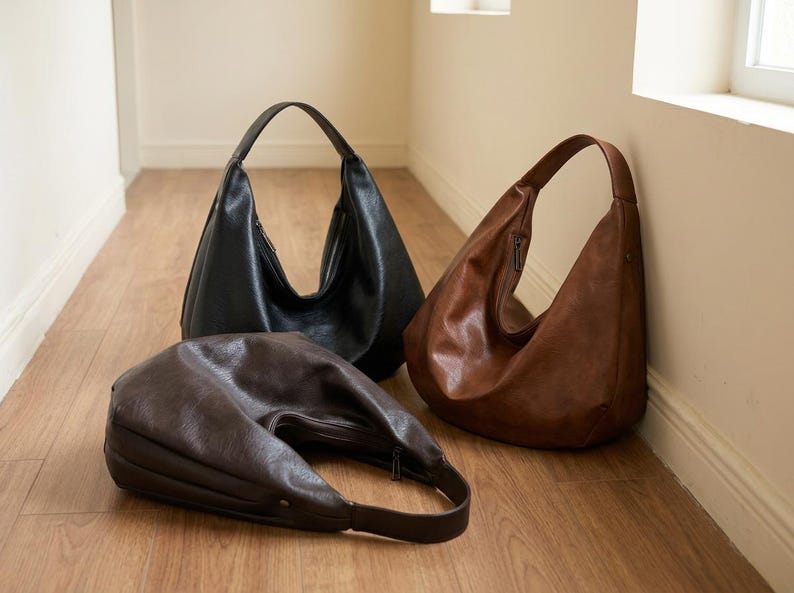 Op de afbeelding: Drie leren hobo-tassen in zwart, bruin en donkerbruin worden getoond op een houten vloer. De tassen hebben een halvemaanvorm, een enkele schouderriem en een ritssluiting. De tassen zijn leeg en staan rechtop.
