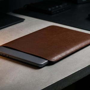 Puede incluir: Una funda de cuero marrón para portátil con un portátil parcialmente insertado. La funda es rectangular con esquinas redondeadas y un borde cosido. El portátil es plateado y encaja perfectamente en la funda. La funda está sobre una superficie gris.