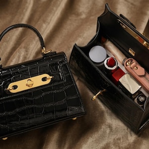 May include: Two black, crocodile-textured handbags. One is closed, featuring a gold-tone clasp and handle. The other is open, revealing cosmetics like lipstick and a small pink zippered pouch. The bags sit on a textured, brown surface.
