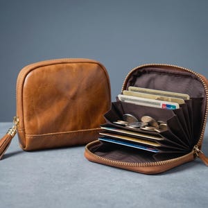 Pode incluir: Uma carteira de couro castanho e uma bolsa de moedas a condizer são exibidas. A carteira está aberta, revelando vários cartões de crédito e moedas. A bolsa de moedas tem um puxador de zíper com borla.