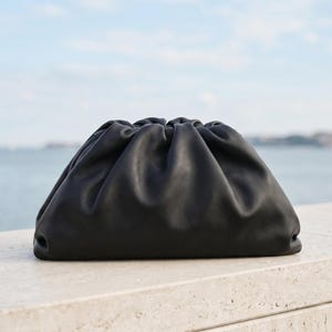 以下が含まれることがあります： 黒い革のクラッチバッグ。上部はギャザーになっており、明るい色の石の表面に置かれています。バッグの柔らかく丸みを帯びた形は、水面と水色の空を背景にしています。