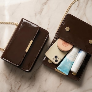 May include: Two brown patent leather shoulder bags with gold chain straps. One bag is closed, the other is open, revealing a phone, makeup, and skincare products. The bags are on a marble surface.