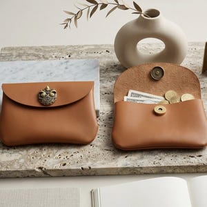 May include: Two tan leather pouches with rounded flaps. One pouch is closed with a decorative antique brass owl emblem, while the other is open, revealing a magnetic snap closure, paper currency, and several gold-toned coins inside the main compartment.