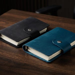 May include: Two leather-bound notebooks, one black and one teal, are displayed on a wooden surface. Each notebook has a flap closure with a silver snap. The pages are white and the leather appears textured.