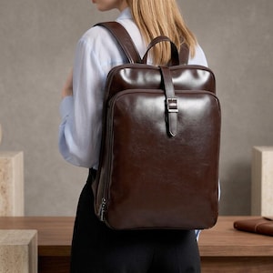 Puede incluir: Una mochila de cuero marrón oscuro con un diseño rectangular. La mochila tiene un asa superior, correas ajustables y un cierre de hebilla. La bolsa se lleva en la espalda.