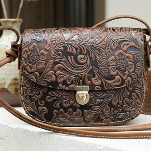 Pode incluir: Bolsa transversal de couro castanho com um design floral em relevo. A bolsa apresenta um fecho de aba com um fecho dourado e uma alça longa e ajustável. A bolsa é do estilo alforge, com costuras brancas.