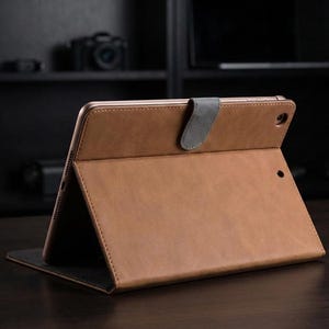 May include: A brown leather tablet case with a grey magnetic clasp. The case is open, showing the tablet screen. The case is made of a textured material and is on a dark wooden surface.