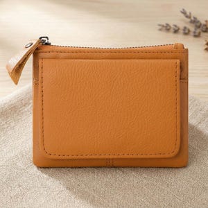 May include: A rectangular mustard yellow leather wallet featuring a top zipper closure with a matching leather pull tab and a front exterior pocket with visible stitching along the edges. The wallet has a smooth, consistent texture and a clean design.
