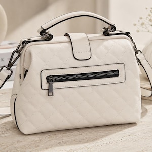 May include: Cream-coloured handbag with a quilted pattern. The bag features a top handle, a detachable shoulder strap, and a zipped pocket on the back. The hardware is black, and the bag is on a light-coloured surface.