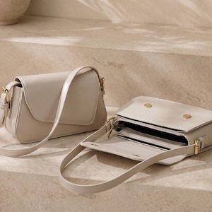 May include: Two beige shoulder bags with smooth surfaces and gold-toned hardware. One bag is closed with a flap, while the other is open, revealing an interior compartment with a central divider and two gold-toned magnetic snap closures.