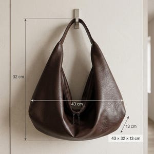 Op de afbeelding: Een donkerbruine leren hobo-tas, 43 cm breed, 32 cm hoog en 13 cm diep, hangt aan een metalen haak. De tas heeft een ritssluiting en een enkele schouderriem. De afmetingen van de tas worden op de afbeelding weergegeven.
