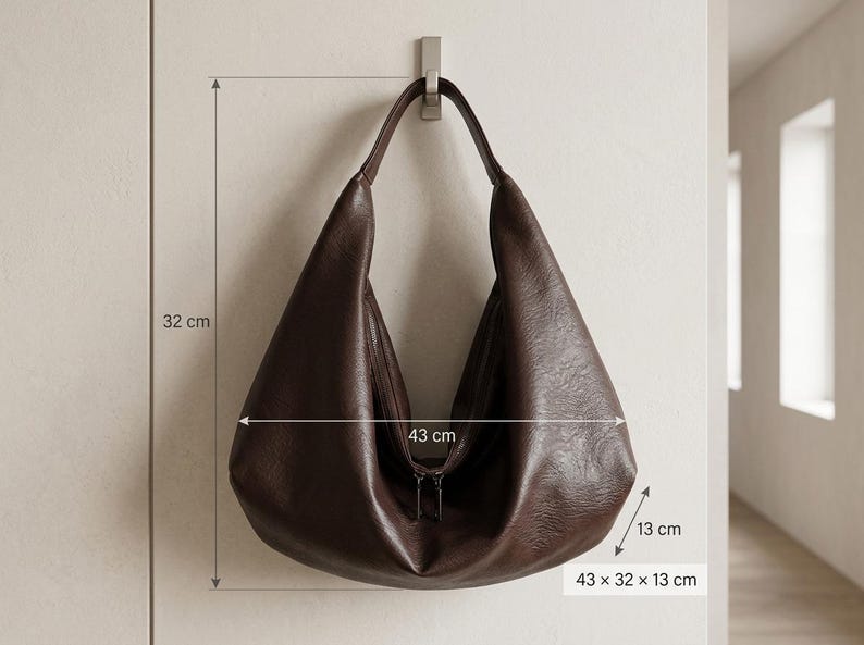 Op de afbeelding: Een donkerbruine leren hobo-tas, 43 cm breed, 32 cm hoog en 13 cm diep, hangt aan een metalen haak. De tas heeft een ritssluiting en een enkele schouderriem. De afmetingen van de tas worden op de afbeelding weergegeven.