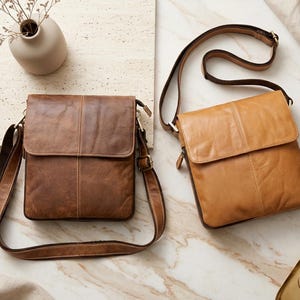 May include: Two rectangular crossbody bags with flap closures and adjustable shoulder straps. One bag is a dark brown, and the other is a lighter tan. Both bags feature vertical stitching details on the front flap and metal hardware accents.