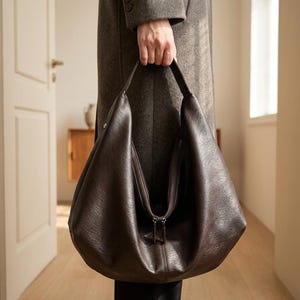 Op de afbeelding: Een donkerbruine hobo-tas met ritssluiting. De tas wordt vastgehouden door een persoon die een lange, grijze jas draagt. De tas heeft een zacht, nonchalant ontwerp en een enkele schouderriem. De achtergrond omvat een deuropening en een glimp van een kamer.