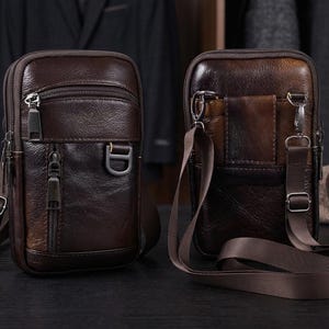 May include: Two brown leather crossbody bags with adjustable straps and multiple zipped compartments. The bags have a rectangular shape and a vintage look. They are displayed on a dark surface, ideal for carrying essentials.