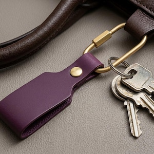 May include: A close-up of a purple leather key fob with a gold-colored carabiner and a small brass button. A set of keys hangs from the carabiner. The fob is attached to a brown leather bag handle.
