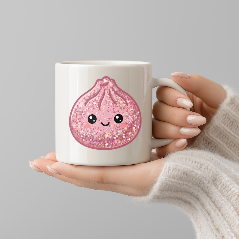 May include: White ceramic mug featuring a pink dumpling illustration with a smiling face and glitter accents. The mug has a curved handle and is held by a person wearing a cream-colored sweater. The design is whimsical and food-themed.