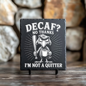 Puede incluir: Posavasos de pizarra negra con el texto "DECAF? NO THANKS I'M NOT A QUITTER." Un p&aacute;jaro de dibujos animados con expresi&oacute;n enfadada sostiene un bate de b&eacute;isbol y una taza de caf&eacute;. El posavasos se muestra sobre un soporte negro.