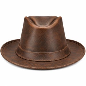 Könnte beinhalten: Ein brauner Fedora-Hut mit einer abgenutzten, verwitterten Textur. Der Hut verfügt über eine eingedrückte Krone, eine strukturierte Krempe mit genähten Kanten und ein passendes Band, das die Basis der Krone umschließt, in einem einheitlichen Dunkelbraun.