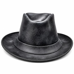 Może przedstawiać: Czarny kapelusz typu fedora o postarzałej, wyblakłej fakturze. Kapelusz posiada wgniecioną główkę, usztywnione rondo z przeszytym brzegiem oraz szeroką, dopasowaną taśmę wokół podstawy główki. Materiał wygląda na gładki z subtelnymi różnicami.