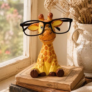 May include: A hand-carved wooden giraffe figurine with a yellow and brown spotted pattern, featuring a smiling expression and large ears. The giraffe is wearing a pair of black-rimmed eyeglasses across its face, resting on its snout.