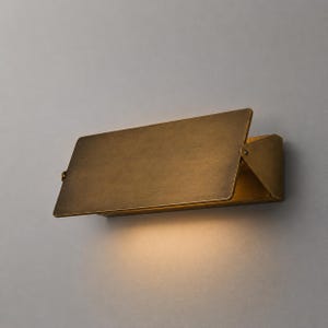 May include: A rectangular wall sconce with a brushed brass finish. The fixture features a flat, angled front panel that extends outward from a triangular side mounting bracket, casting a warm, downward glow against the wall surface behind it.