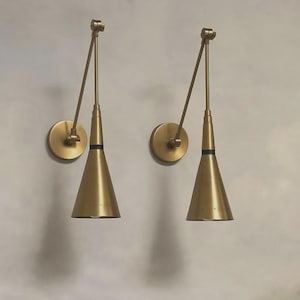 May include: Two brass wall sconces with a cone-shaped shade and adjustable arm. The sconces have a circular base and a black band near the top of the shade. The design is modern and minimalist.
