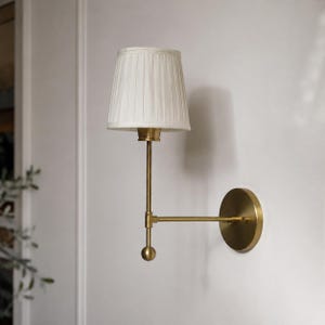 May include: A wall-mounted lamp featuring a brushed brass finish arm and a circular wall plate. The fixture includes a vertical rod ending in a spherical finial, supporting a white, pleated, tapered fabric lampshade that directs light downwards.
