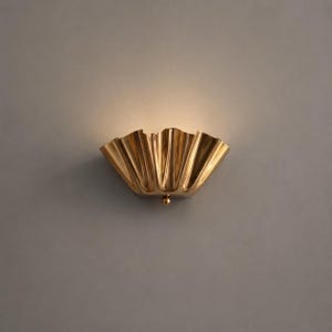 May include: A gold-toned wall sconce with a fluted, shell-like design. The fixture features a metallic, reflective surface with deep vertical folds and a small, spherical knob at the bottom centre. It emits a soft, warm glow from the top opening.