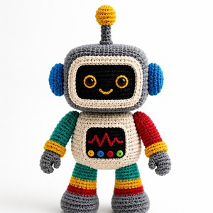 May include: A crocheted robot toy featuring a gray head with a yellow antenna, blue ear pieces, and a black face with yellow-rimmed eyes and a smile. The body is cream-colored with a black chest panel, red zigzag stitching, and colorful buttons.