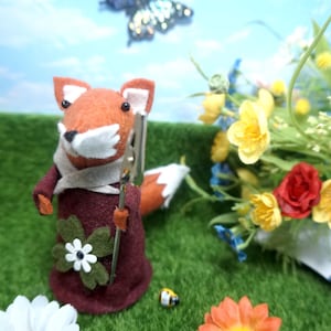 May include: A felt fox figurine with orange fur, a white muzzle, and a burgundy dress, holding a silver tool. A butterfly flies in the background. The scene includes flowers and a small bee figurine on a green surface.