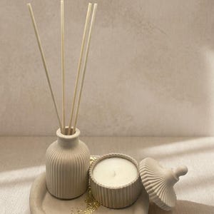 May include: A beige decorative set comprising a small vase with reeds, a candle, and a decorative lid, presented on a round tray. The tray is embellished with gold leaf accents. Ideal for home decor.