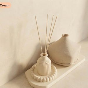 May include: A cream-coloured reed diffuser set featuring a ribbed vase with reeds, a decorative tray, and a geometric vase. The items are arranged on a cream-coloured surface, creating a minimalist aesthetic. The theme is home fragrance and decor.