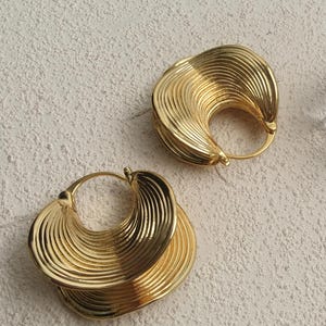 May include: A pair of gold-toned hoop earrings featuring a distinct fan-like shape with concentric, raised ribbed lines radiating from the centre. The earrings have a polished metallic finish and a circular opening, accompanied by a clear acrylic cube.