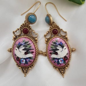May include: A pair of gold-toned drop earrings featuring an oval mosaic inlay of a white bird surrounded by blue and red flowers on a pink background. Each earring includes a small round blue stone at the top and a dark red stone accent.