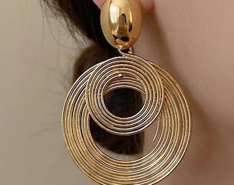 May include: A gold-toned earring featuring a smooth, polished oval stud at the top, connected to two concentric circular hoops below. The hoops are constructed from thin, coiled wire, creating a textured, layered appearance in a bright metallic finish.