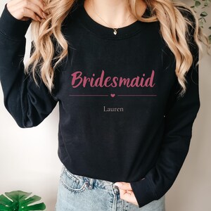 May include: A black long-sleeve crewneck sweatshirt featuring the word Bridesmaid in a dark pink script font, followed by a horizontal line with a small heart in the center and the name Lauren printed in a smaller, simple font below it.