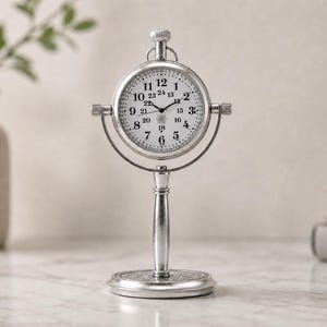 May include: A silver-toned metal desk clock featuring a round face with black Arabic numerals from 1 to 12 and smaller 24-hour markings from 13 to 24. The clock is mounted on a vertical stand with a circular base and a top winding knob.