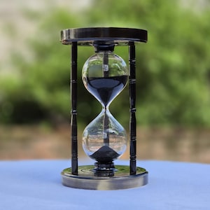 Peut inclure: Un sablier doté d'un cadre en bois noir avec quatre piliers verticaux reliant une base et un sommet circulaires. Le bulbe en verre transparent contient du sable noir fin, dont une partie repose dans la chambre inférieure et le reste au-dessus.