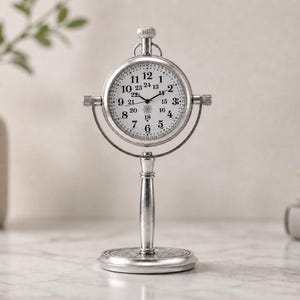May include: A silver-colored desk clock with a round face and a stand. The clock face has black numerals and hands. The clock is supported by a decorative stand with a circular base. The clock measures approximately 6 inches in height.