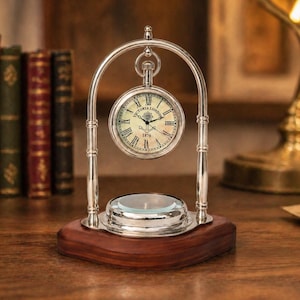 Op de afbeelding: Een zilverkleurige zakhorloge bureauklok met een witte wijzerplaat en Romeinse cijfers. De klok hangt in een decoratief zilveren frame en staat op een houten voet. De klok leest "Victoria London 1876".