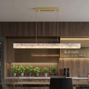 Könnte beinhalten: Eine moderne rechteckige Pendelleuchte mit goldfarbener Deckenhalterung und einem langen, mattierten Glasschirm. Das Licht hängt über einer Küchenzeile mit Topfpflanzen und einem dunklen Holzschrank.