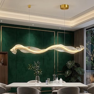 May include: A contemporary, gold-coloured, undulating chandelier suspended above a dining table. The light fitting has a ribbon-like design with integrated lighting. The room features a dark green wall with gold trim and a marble-topped table.