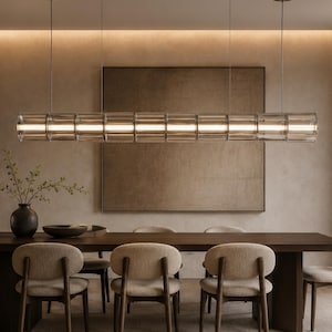 Könnte beinhalten: Ein modernes Esszimmer mit einem langen, rechteckigen Holztisch und sechs beige gepolsterten Stühlen. Über dem Tisch hängt eine lineare Pendelleuchte aus klarem Glas und Metall mit interner Beleuchtung. Eine dunkle Vase und eine Schüssel stehen auf dem Tisch.