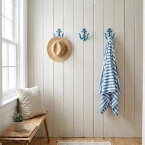May include: Three blue anchor-shaped wall hooks mounted on a vertical white paneled wall. A straw hat hangs on the left hook, and a blue and white striped towel hangs on the right hook. A wooden bench with a beige pillow and a small potted plant sits below.