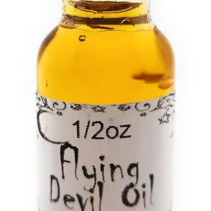 Puede incluir: Una pequeña botella de vidrio llena de líquido dorado, etiquetada como "Flying Devil Oil" con "1/2oz" en la etiqueta. La botella tiene una tapa negra y una etiqueta blanca con texto negro. Producto de Terra Traders.